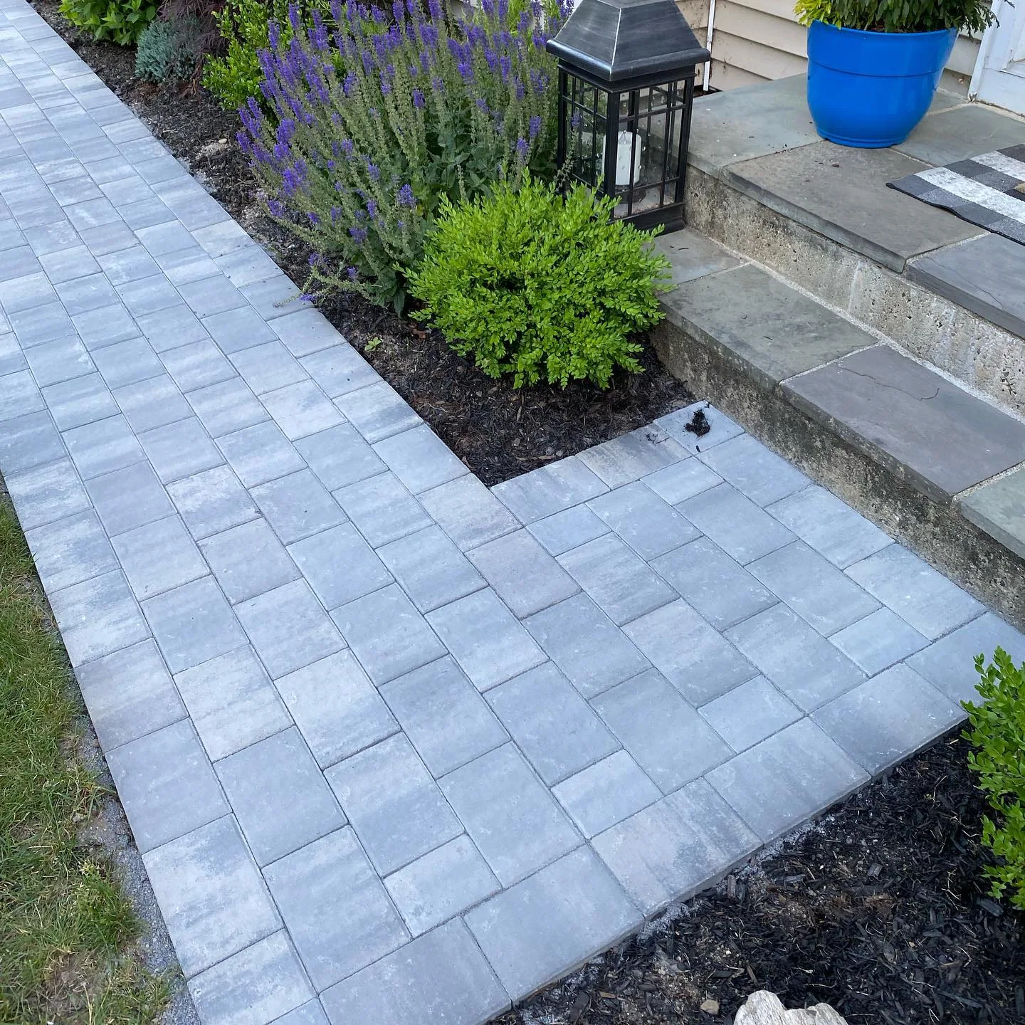 A newly completed modern paver walkway installation featuring gray rectangular stones bordering a landscaped garden with purple flowers.
