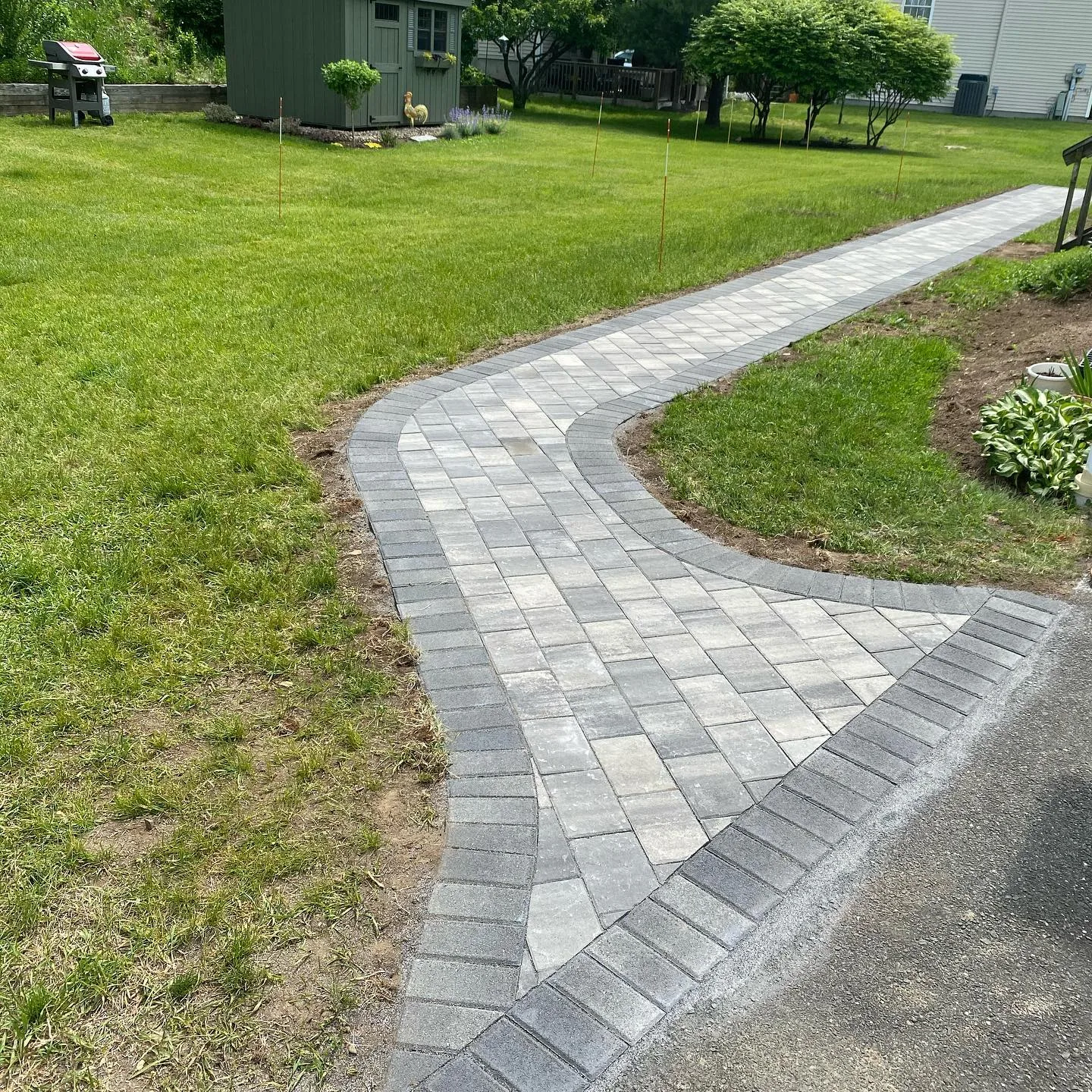 A newly completed interlocking paver walkway installation featuring gray stones and a dark charcoal border winding through a green lawn.