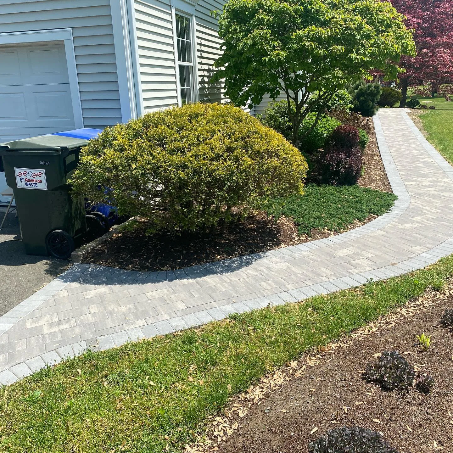 A newly completed paver walkway installation curving around a landscaped garden bed next to a white house.