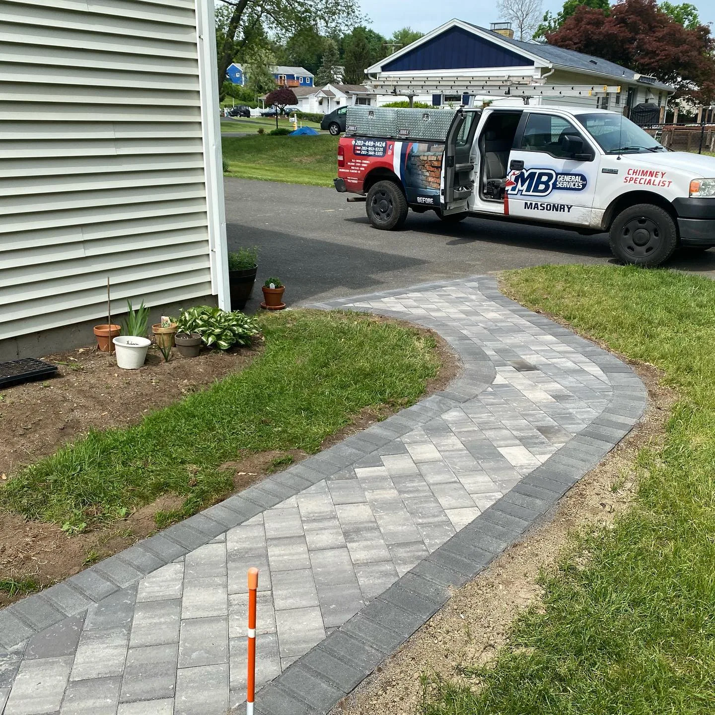 A newly finished paver walkway installation featuring a curved path with light gray stones and a dark border leading to a driveway.