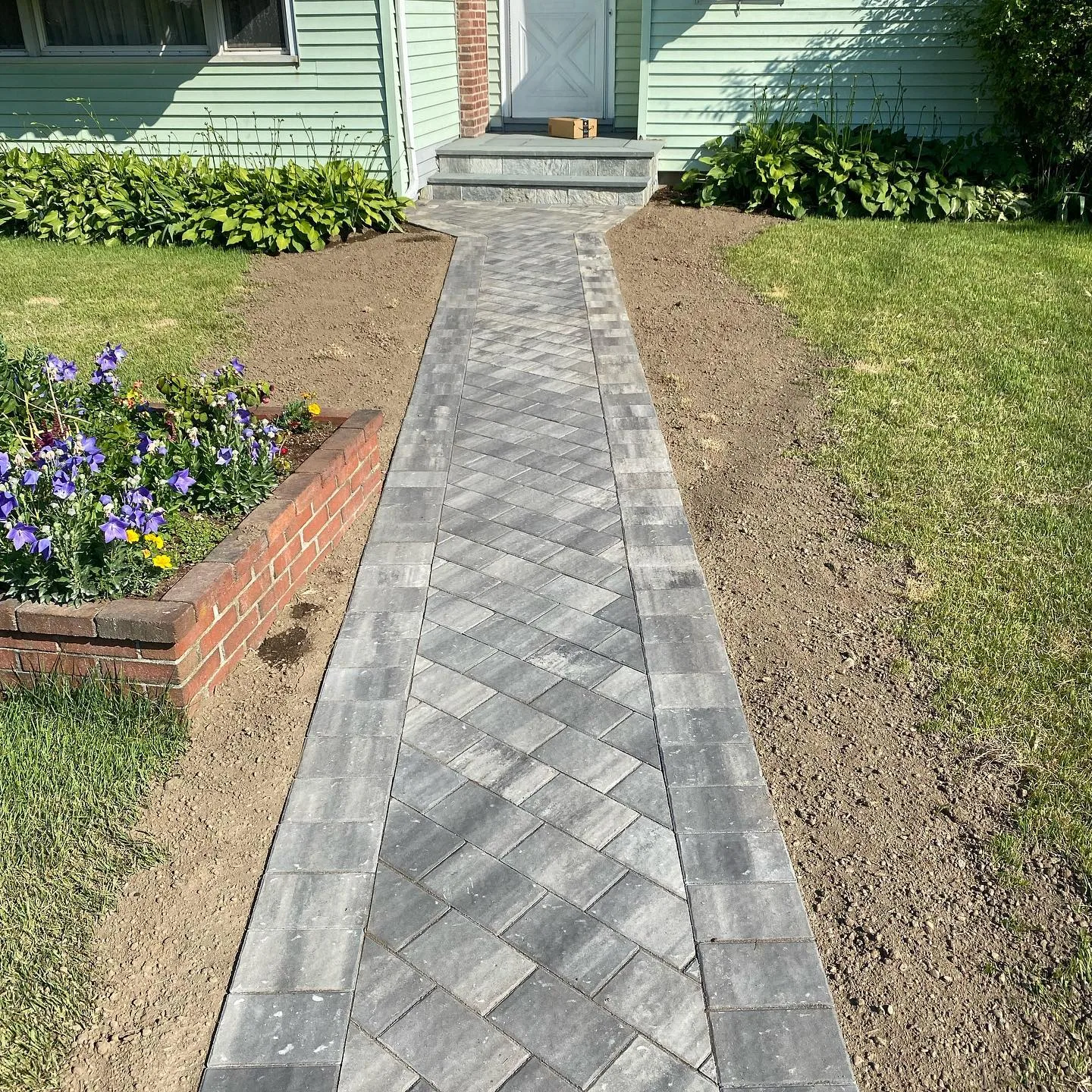 A completed paver walkway installation featuring a gray herringbone pattern leading to a house front door.