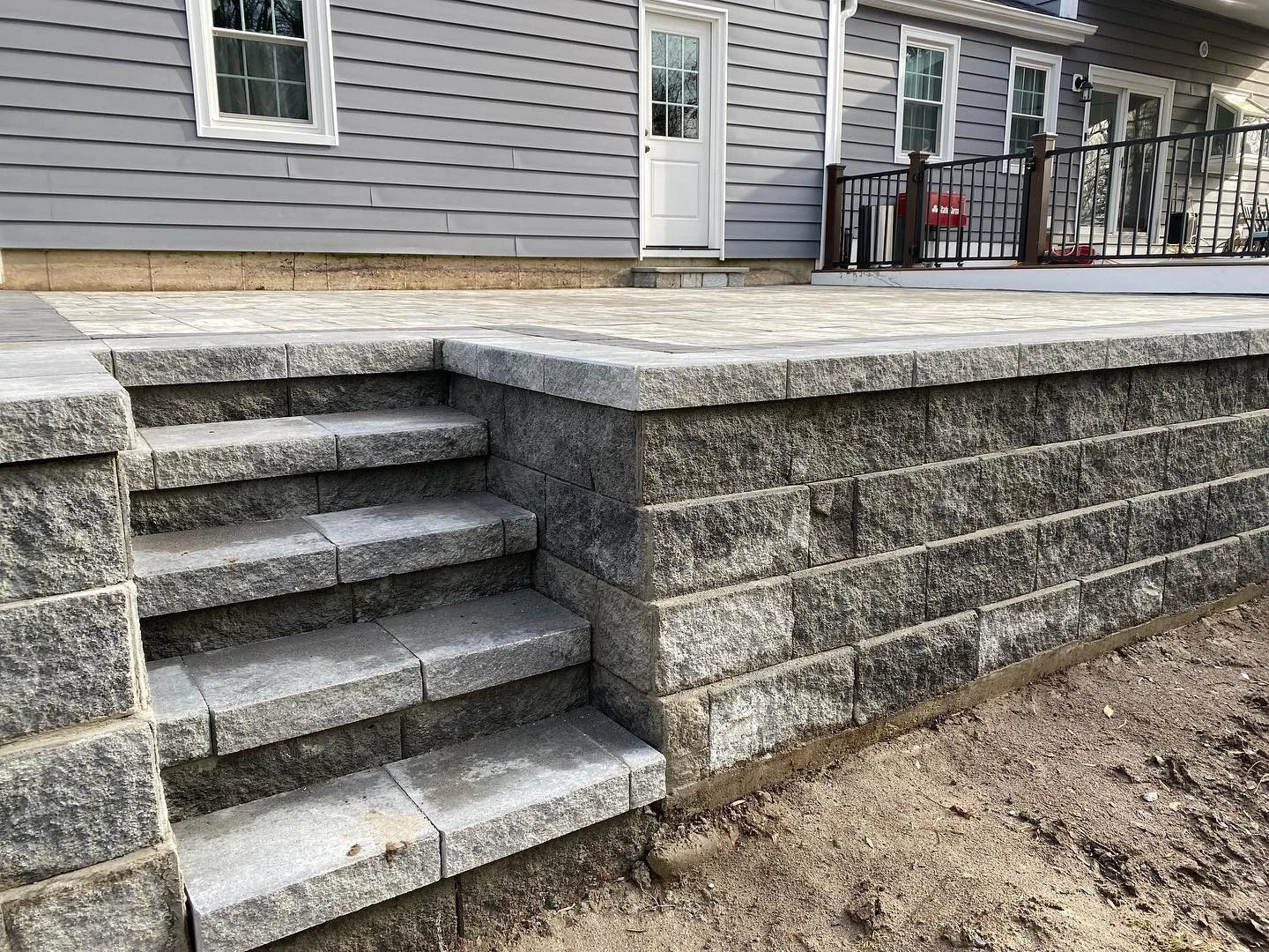 A high-quality raised patio paver installation featuring grey retaining wall blocks and integrated stone steps leading to a modern home.